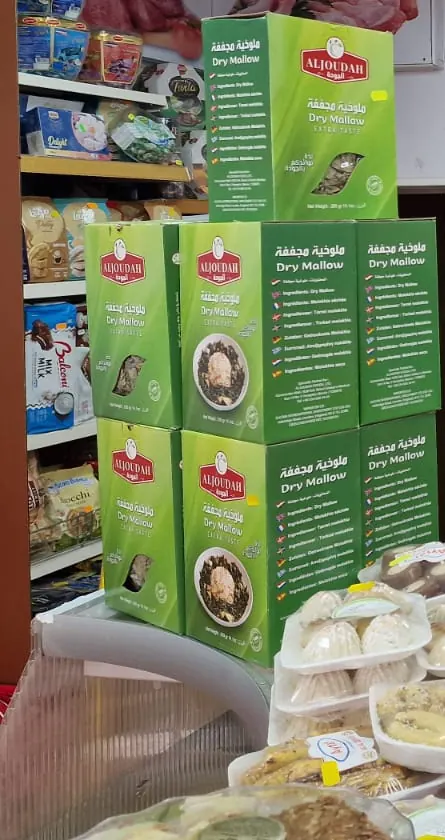 Boxes of Aljoudah dried molokhia stacked above trays of maamoul and pastries.